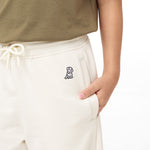 Close-up view of off-white jogger pants featuring a small embroidered dog logo on the side and a drawstring waist, with a child's hand resting in the pocket