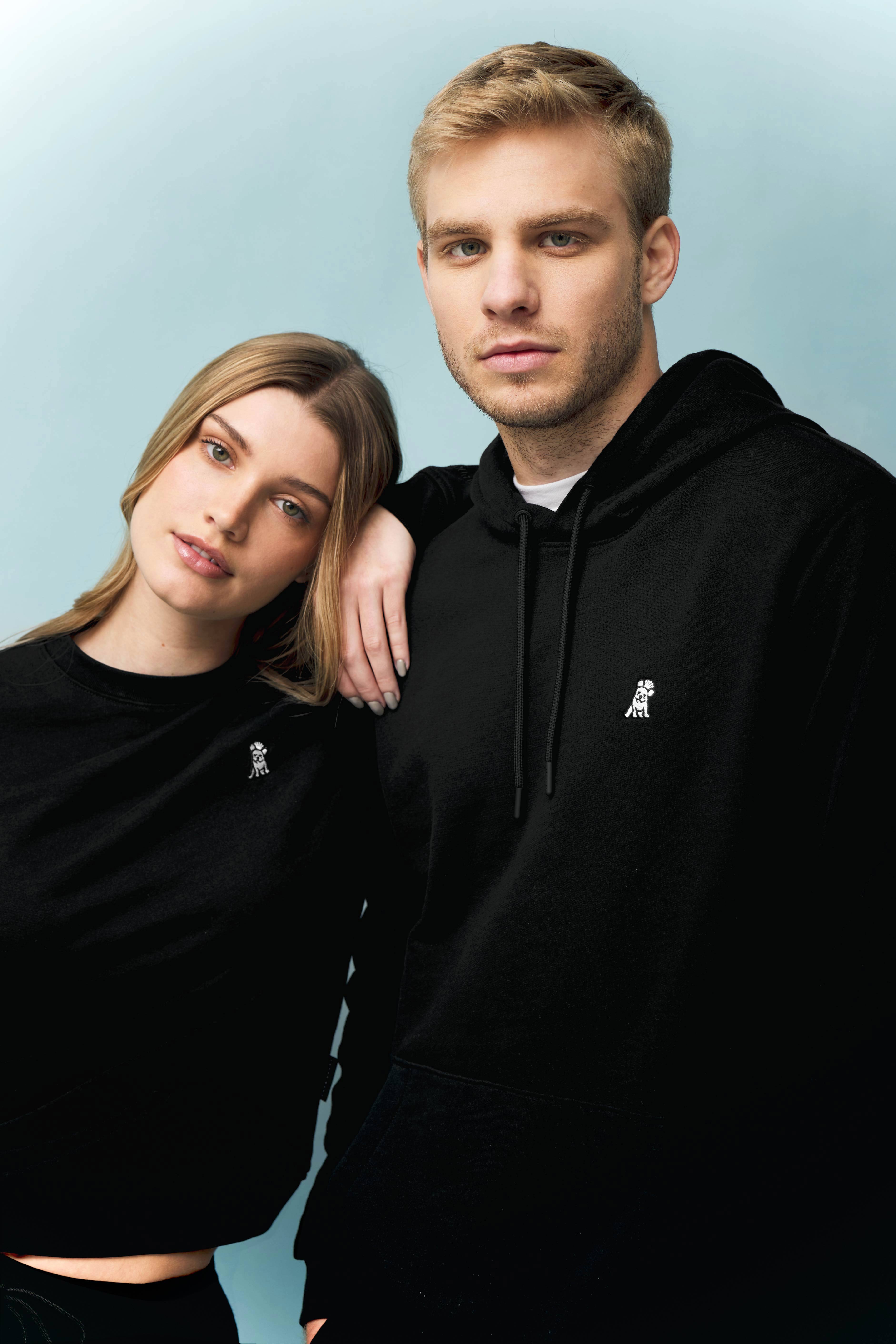 A young man and woman wearing matching black sweatshirts with a small embroidered dog logo on the chest. The woman leans her head on the man's shoulder, and both have a neutral expression. The background is a soft pastel blue.