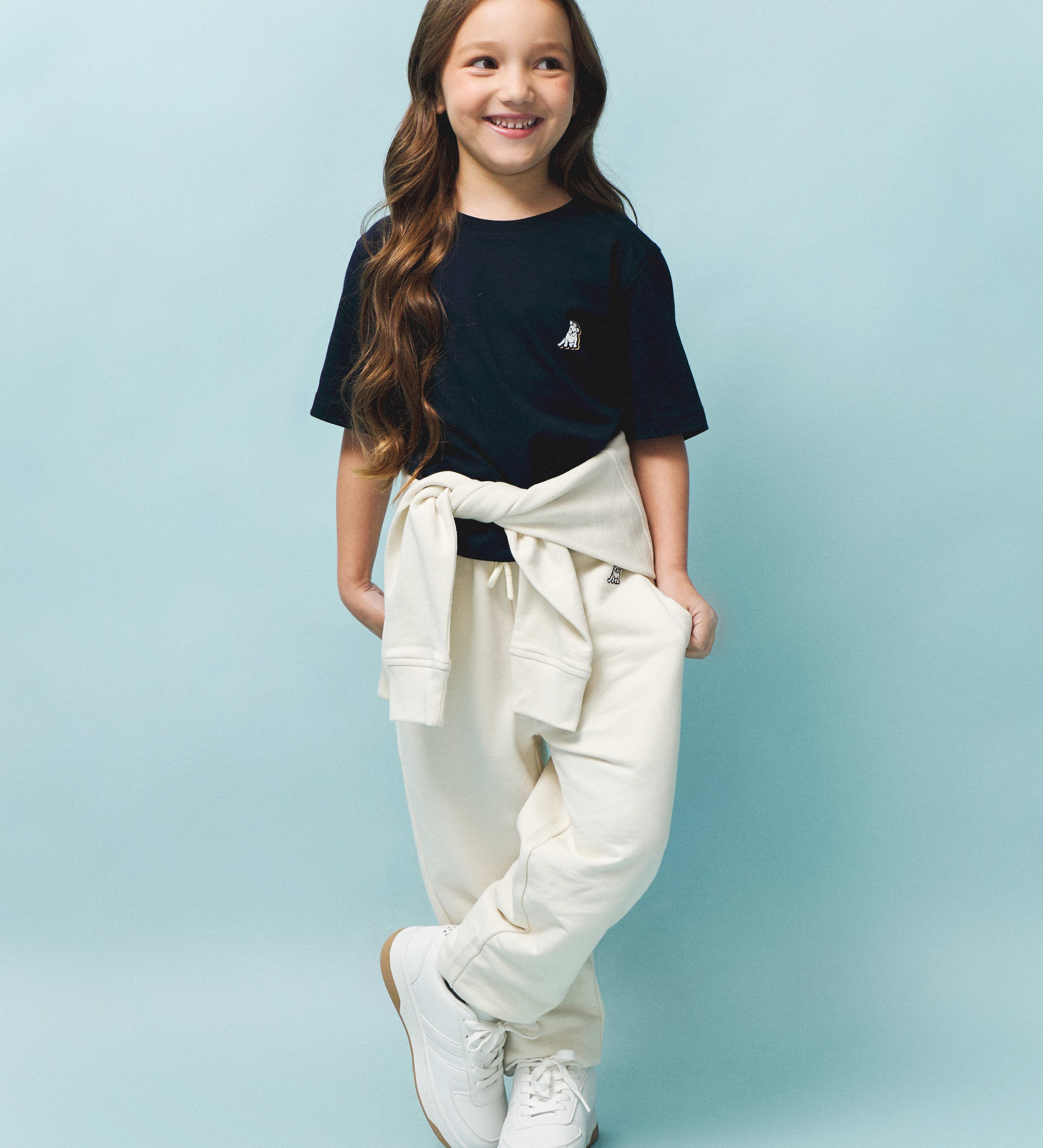 This image features a young girl with long, wavy brown hair, wearing a dark navy t-shirt with a small embroidered dog logo on the left side of the chest. She has light beige jogger pants with a matching beige hoodie tied around her waist. She completes the look with white sneakers. The pastel blue background adds a soft and modern touch, keeping the focus on the stylish and comfortable outfit. 