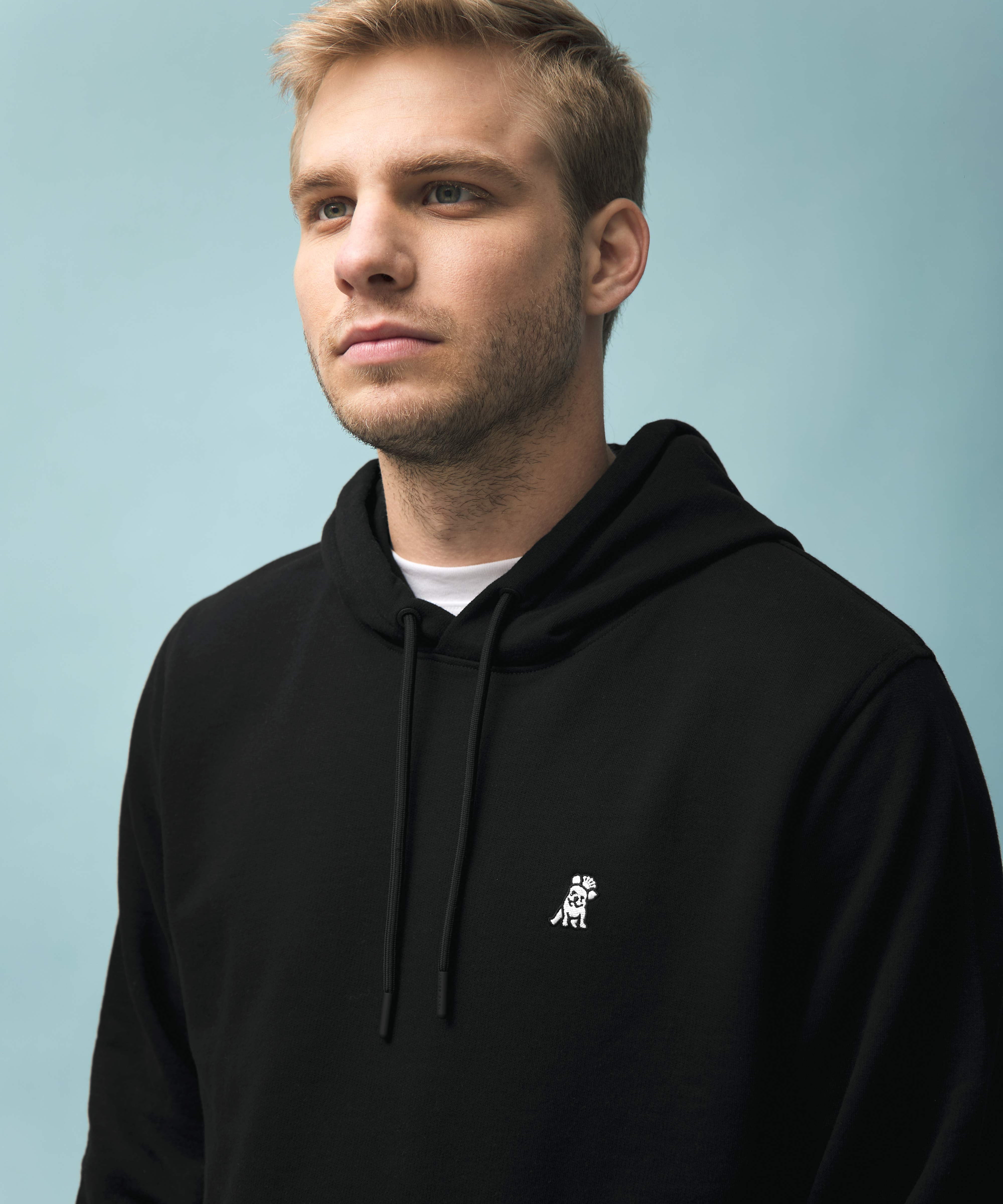 This image features a young man with short blonde hair and light facial stubble, wearing a black hoodie with a small embroidered logo of a dog on the left side of the chest. The hoodie has a drawstring and is layered over a white t-shirt, which is slightly visible at the neckline. The background is a soft pastel blue, providing a clean and modern aesthetic that contrasts with the dark clothing. Let me know if you need any additional details!