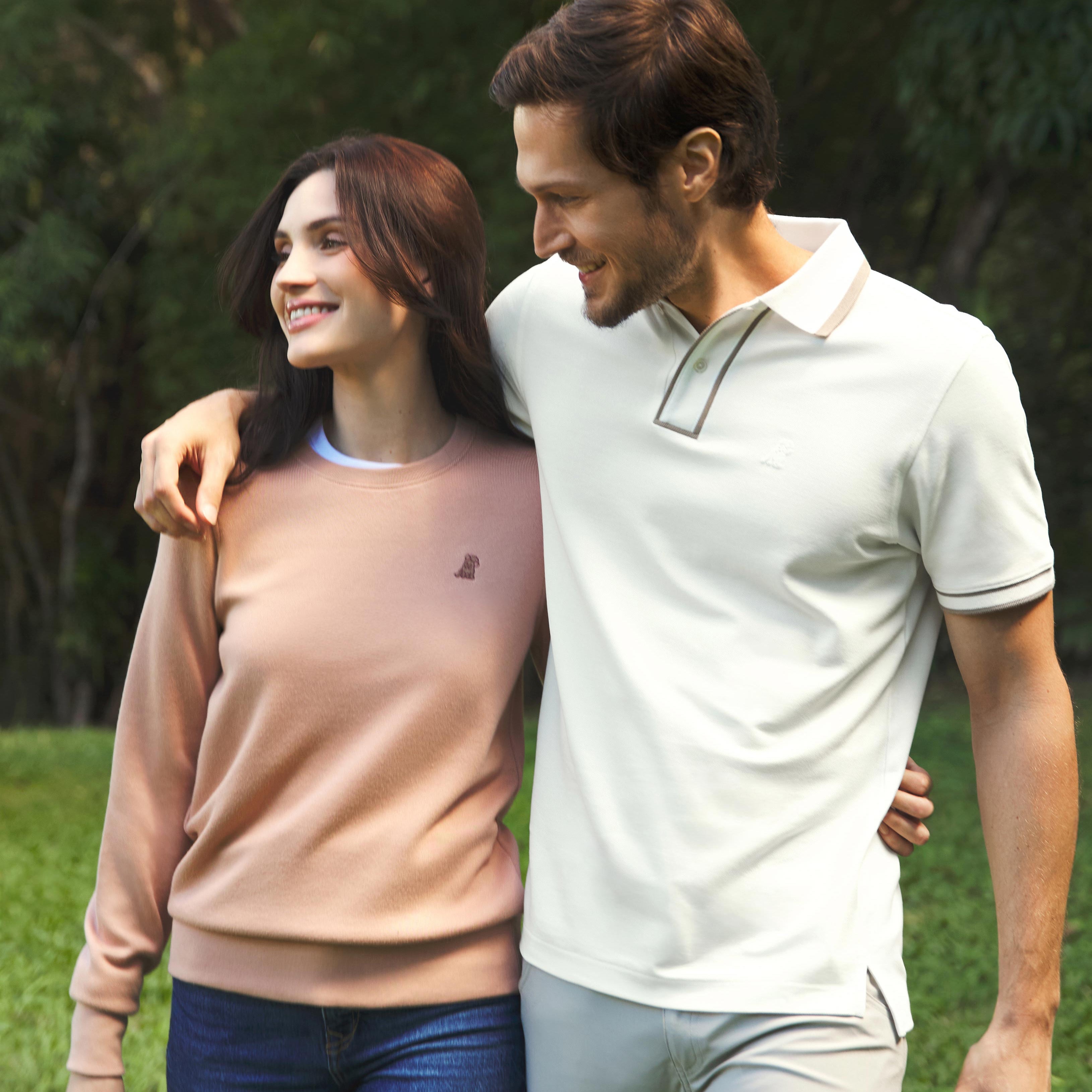 A smiling couple walking together outdoors on a grassy path surrounded by greenery. The woman is wearing a soft pink long-sleeved sweatshirt with an embroidered dog logo and blue jeans, while the man is in a light-colored polo shirt with contrast trim and light-colored pants. They have their arms around each other, sharing a joyful moment.