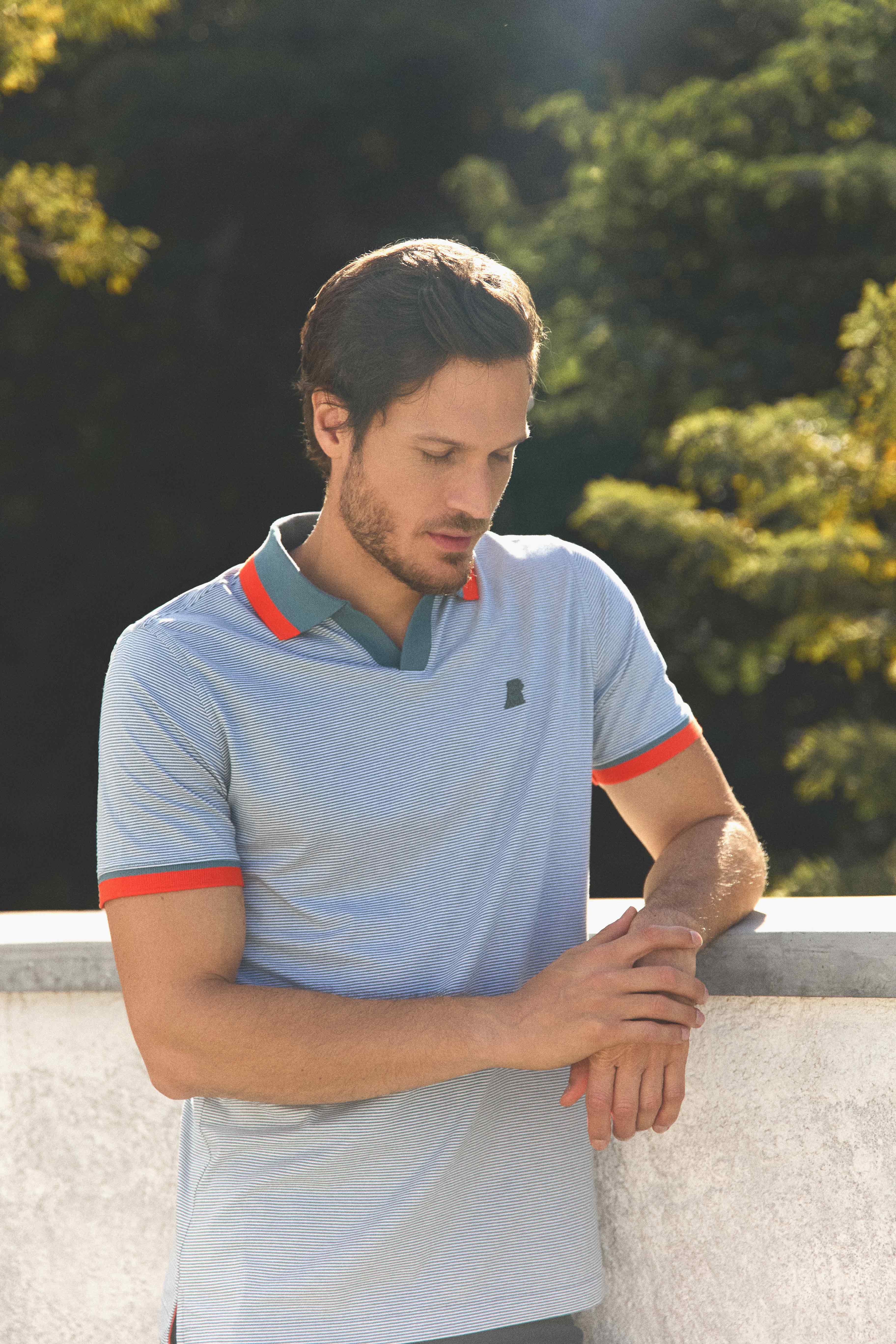 A man with short brown hair and light stubble wearing a light blue striped polo shirt with orange and green collar details. He is looking down at his hands while leaning against a concrete ledge, with a background of green foliage and sunlight filtering through the trees.