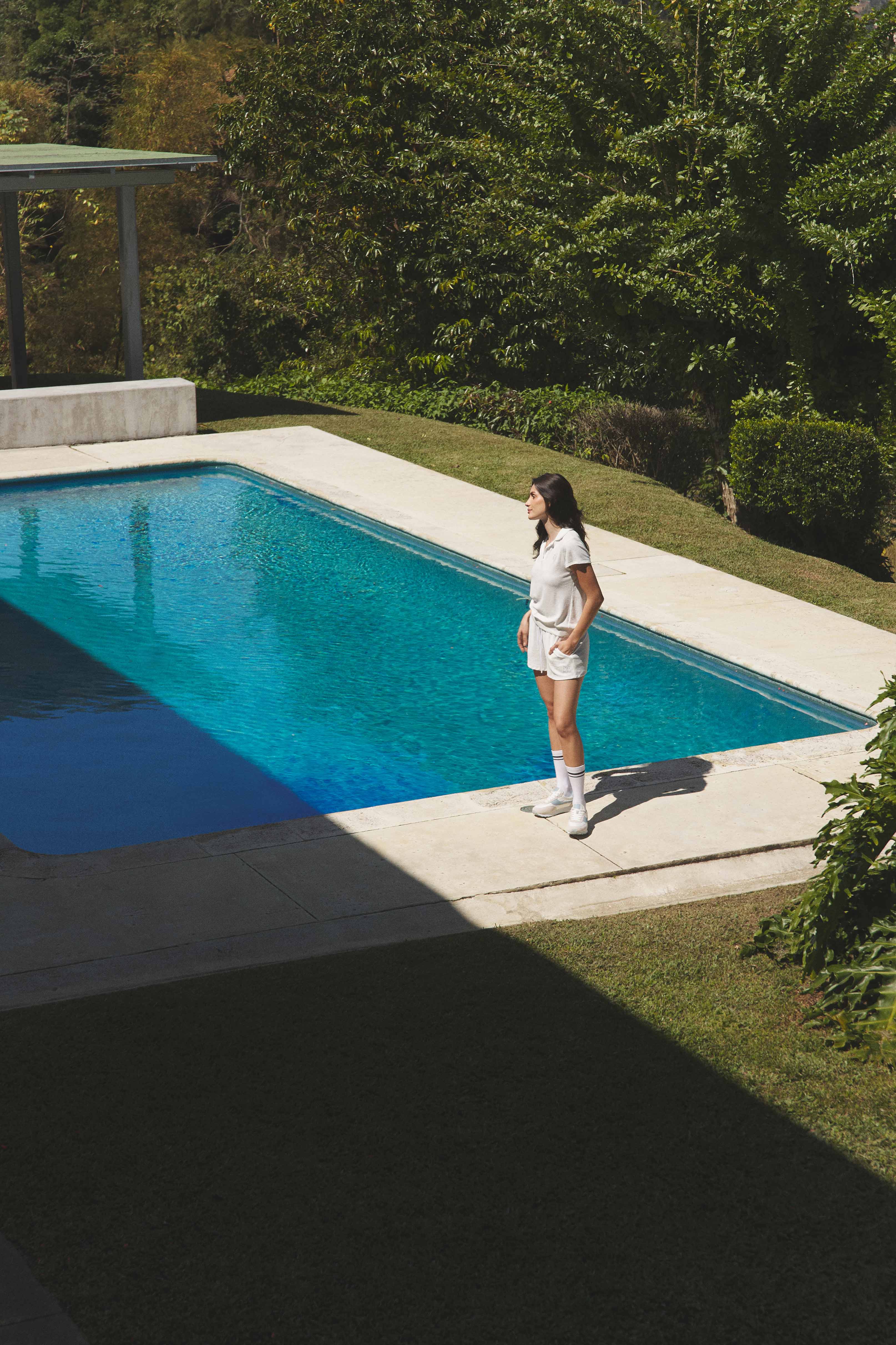 A woman dressed in an all-white sporty outfit, including shorts, a t-shirt, knee-high socks, and sneakers, standing by the edge of a pool. The setting is surrounded by greenery with part of the pool area in shadow, and she gazes off into the distance.