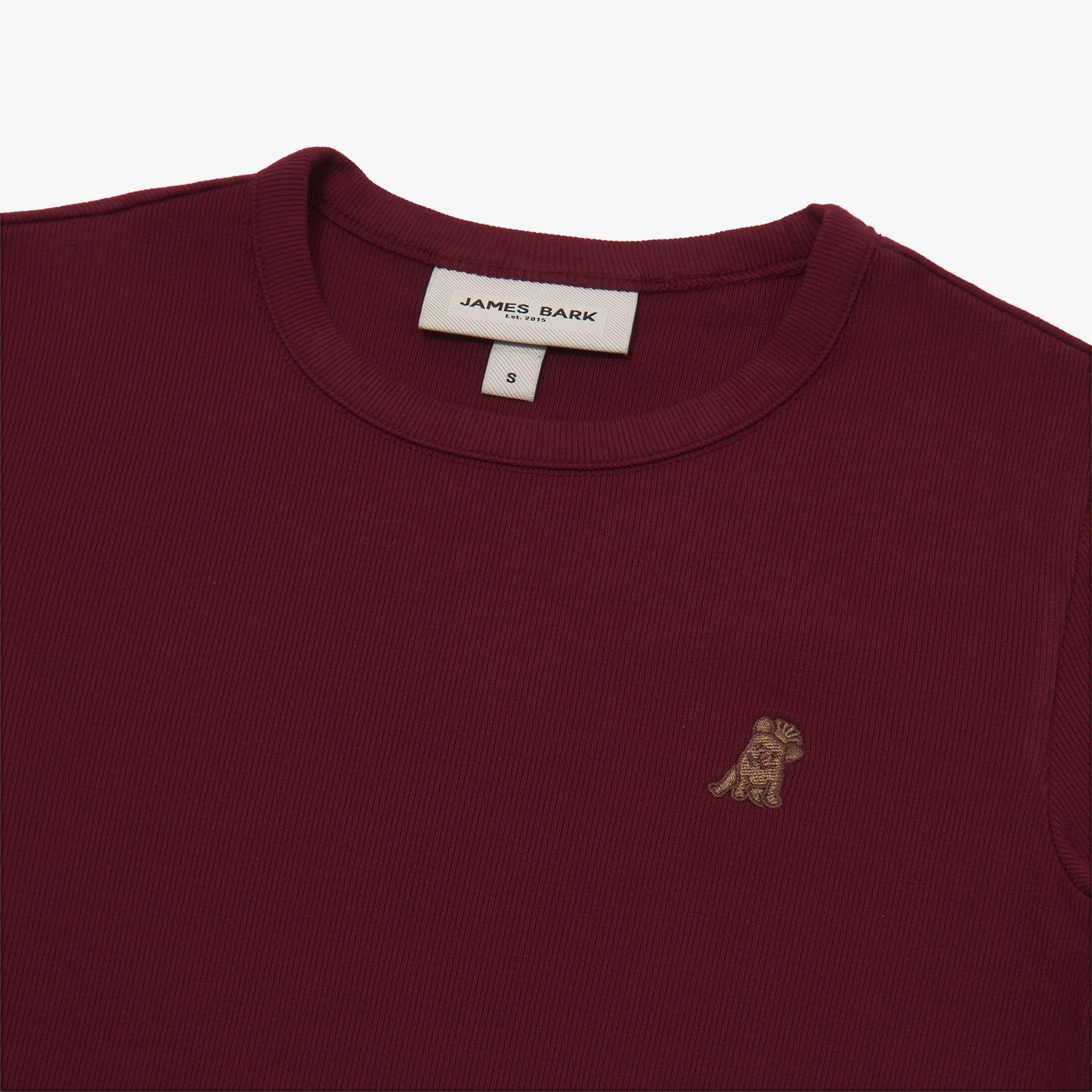 A close-up of a burgundy short-sleeve T-shirt, showing the ribbed crewneck collar and finely knit fabric texture. The white neck label displays the brand name "JAMES BARK" with "Est. 2015" beneath it, along with the size "S." A small embroidered logo of a dog in a slightly darker shade of brown is positioned on the left chest area, subtly contrasting with the fabric. The material appears soft and high-quality.