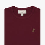 A close-up of a burgundy short-sleeve T-shirt, neatly folded to showcase the ribbed crewneck collar and finely knit fabric texture. The white neck label displays the brand name "JAMES BARK" with "Est. 2015" beneath it, along with the size "S." A small embroidered logo of a dog in a slightly darker shade of brown is positioned on the left chest area. The material appears soft and high-quality