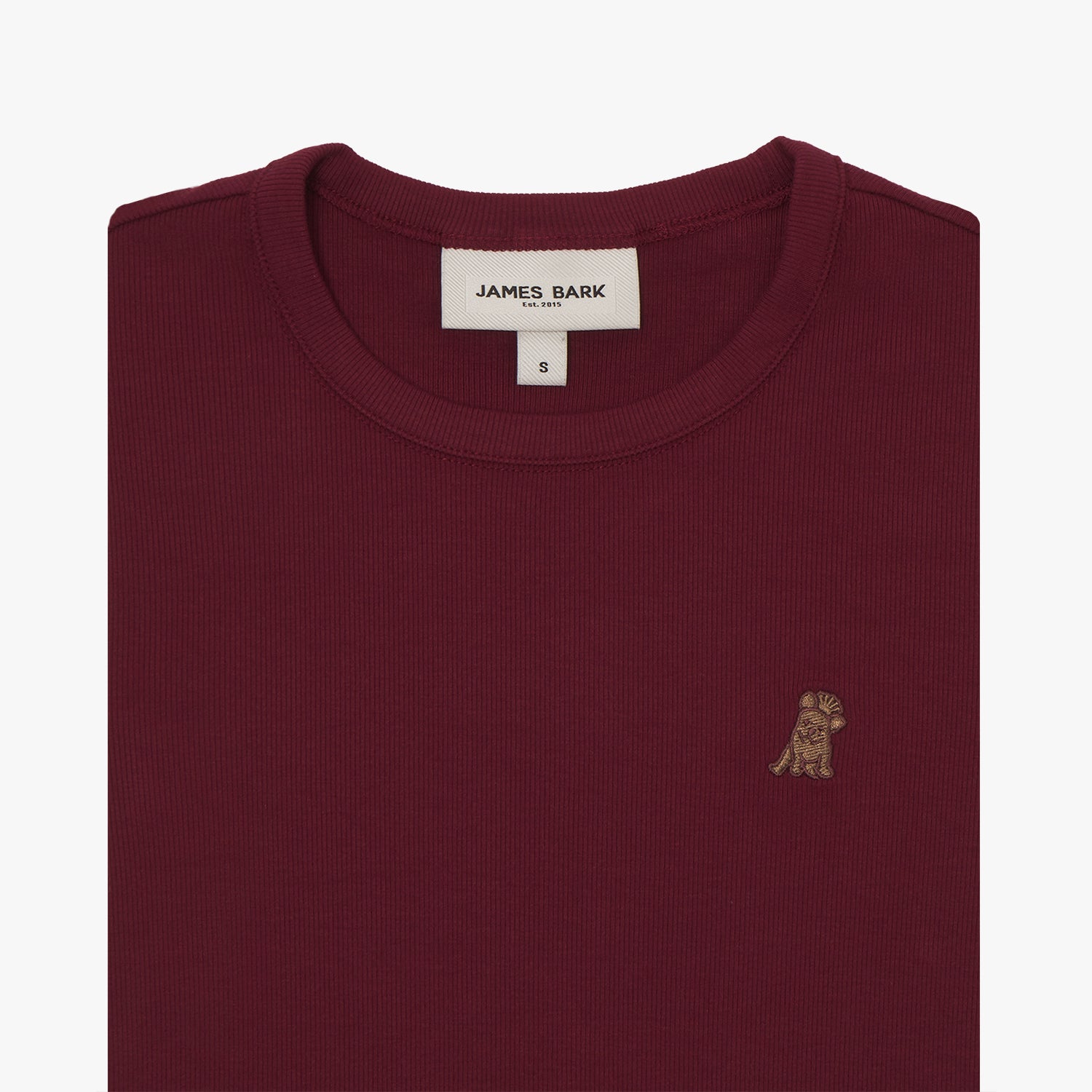 A close-up of a burgundy short-sleeve T-shirt, neatly folded to showcase the ribbed crewneck collar and finely knit fabric texture. The white neck label displays the brand name "JAMES BARK" with "Est. 2015" beneath it, along with the size "S." A small embroidered logo of a dog in a slightly darker shade of brown is positioned on the left chest area. The material appears soft and high-quality