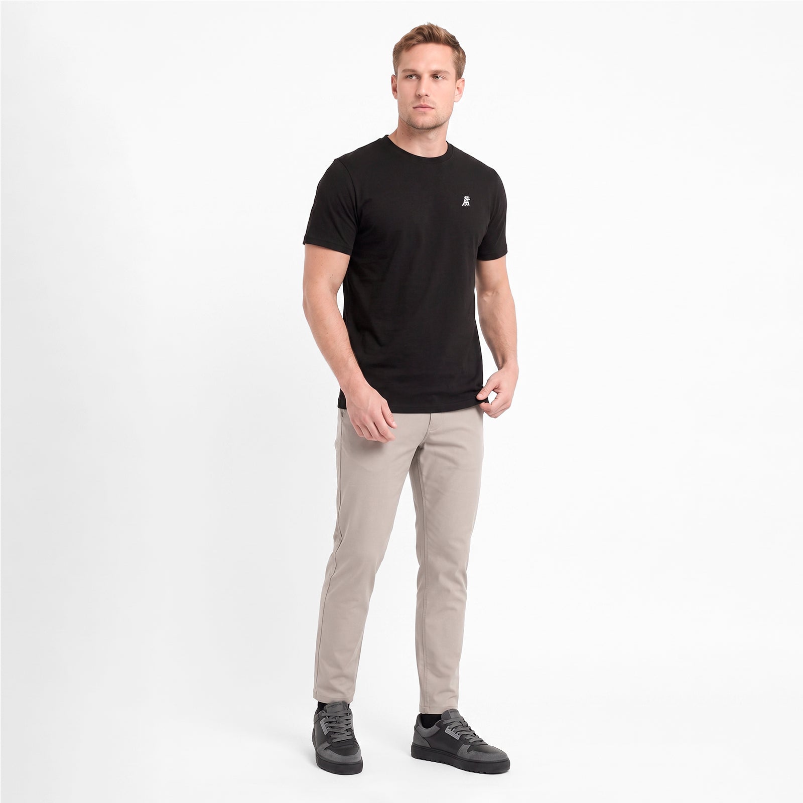 A man wearing black short-sleeve crewneck t-shirt with a small white logo on the left chest, paired with beige trousers