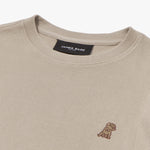 A close-up of a beige crewneck sweatshirt, showing the ribbed collar and fabric texture. The black neck label displays the brand name "JAMES BARK" with "Est. 2015" beneath it. A small embroidered logo of a dog in brown thread is positioned on the left chest area. The material appears to be finely knit and soft