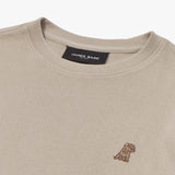 A close-up of a beige crewneck sweatshirt, showing the ribbed collar and fabric texture. The black neck label displays the brand name "JAMES BARK" with "Est. 2015" beneath it. A small embroidered logo of a dog in brown thread is positioned on the left chest area. The material appears to be finely knit and soft