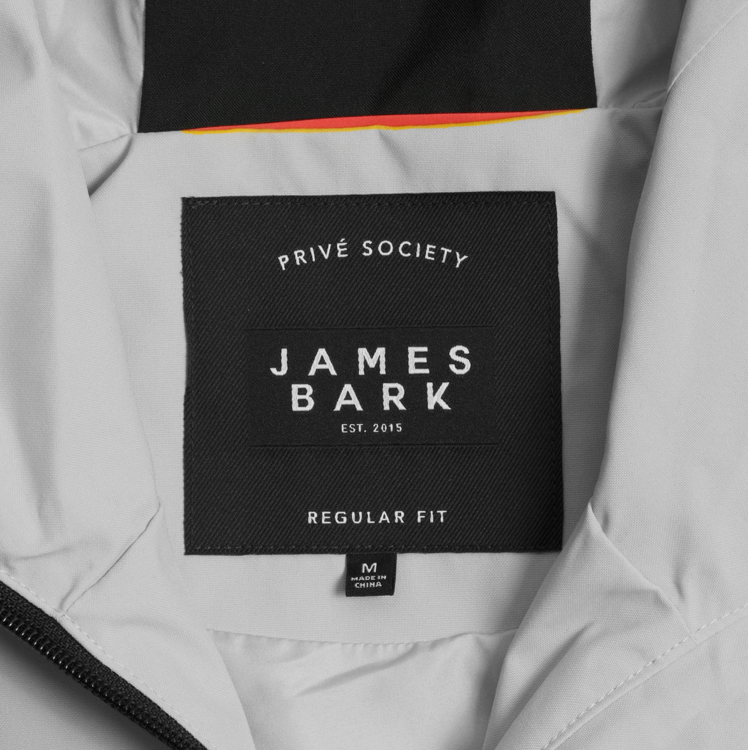 Close-up of the inside label of a light gray jacket. The black label features the text 'PRIVÉ SOCIETY' at the top, with 'JAMES BARK' prominently in the center, followed by 'EST. 2015.' Below, the label reads 'REGULAR FIT,' and a smaller tag beneath indicates the size 'M' and 'Made in China.' A red and yellow accent is visible in the lining above the label.