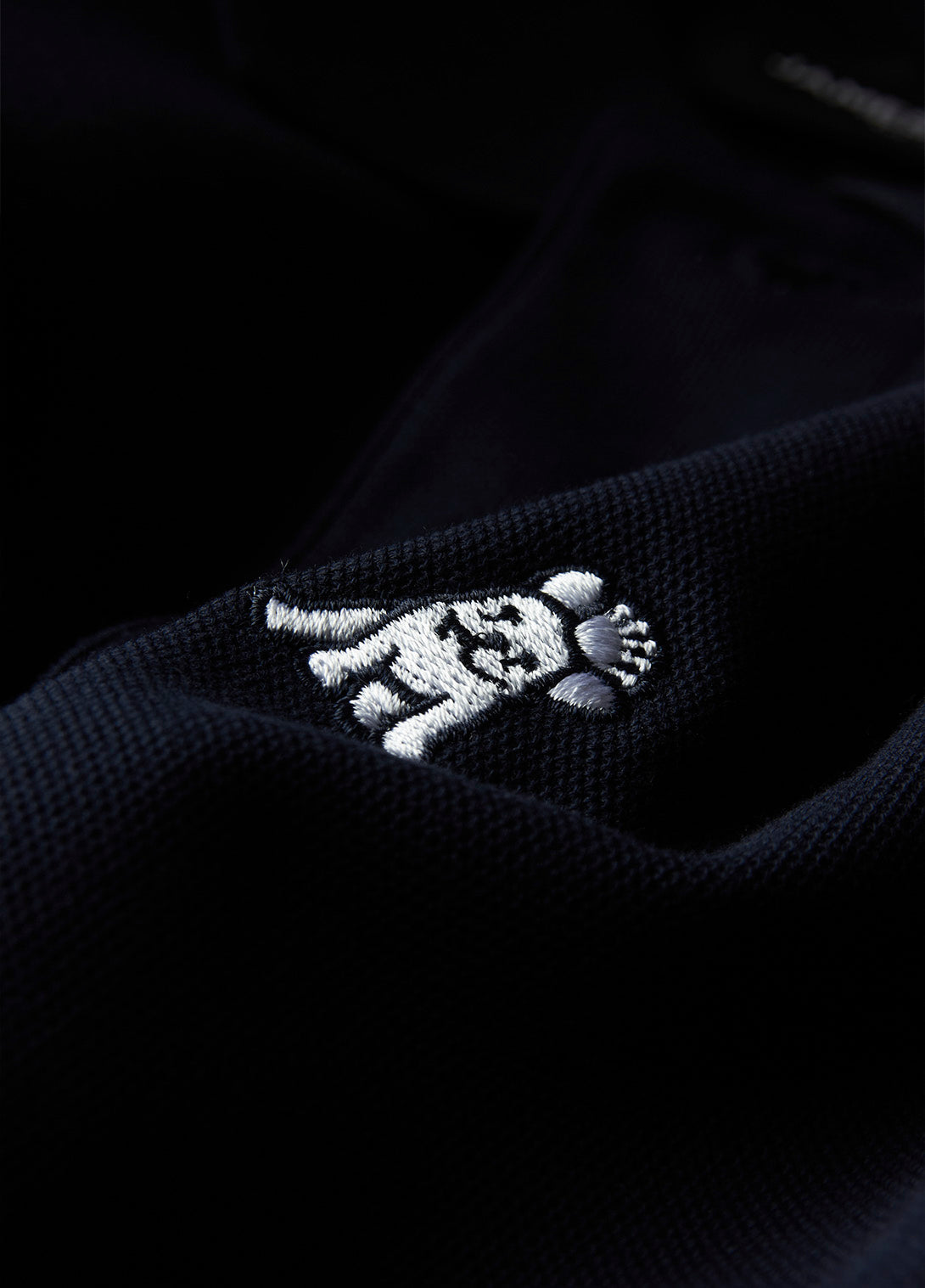 A close-up shows a white embroidered logo of a dog wearing a crown on black or very dark blue textured fabric, likely a polo shirt