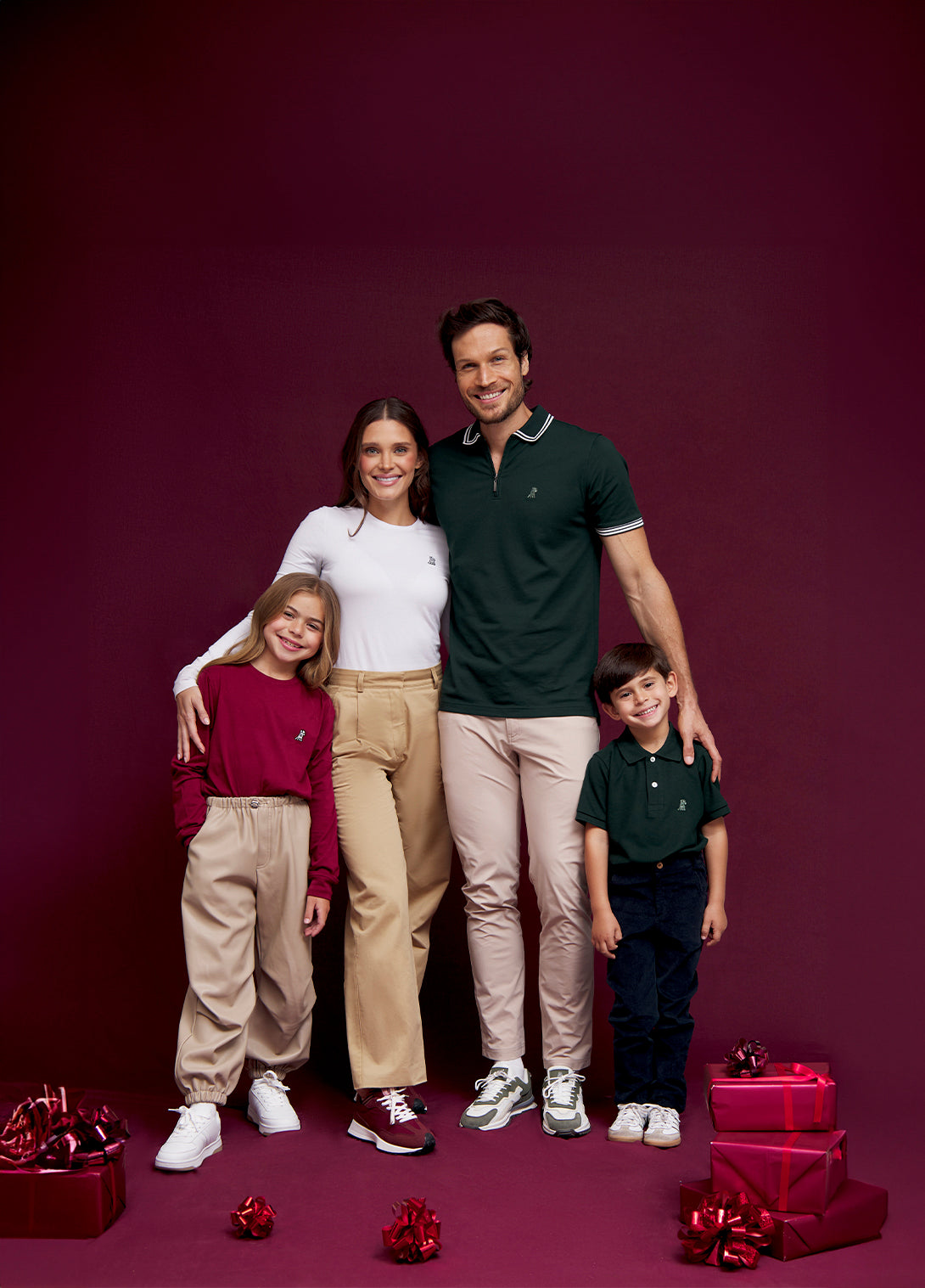 A family of four standing close together, smiling against a burgundy background. The woman in white, the man in a green polo shirt, and the children in maroon and green tops look happy with gift boxes and ribbons at their feet.