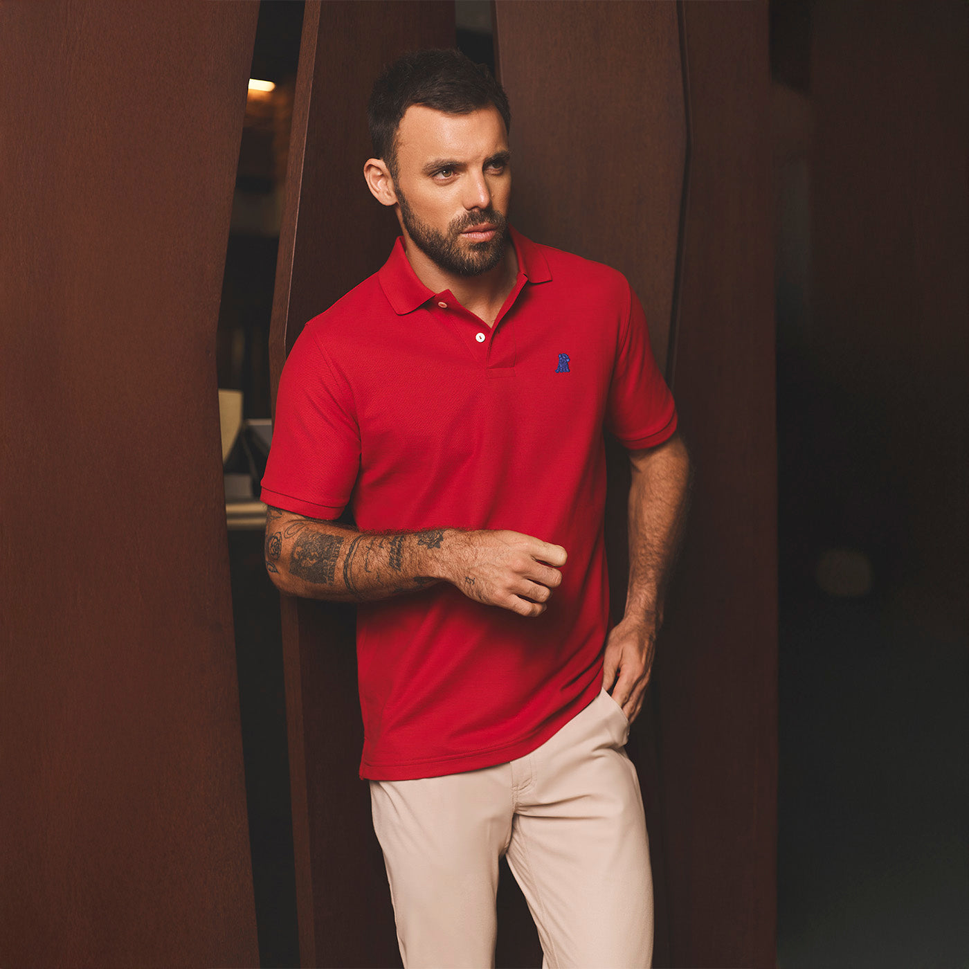 Man wearing a red polo shirt with a logo, standing indoors against a dark background.