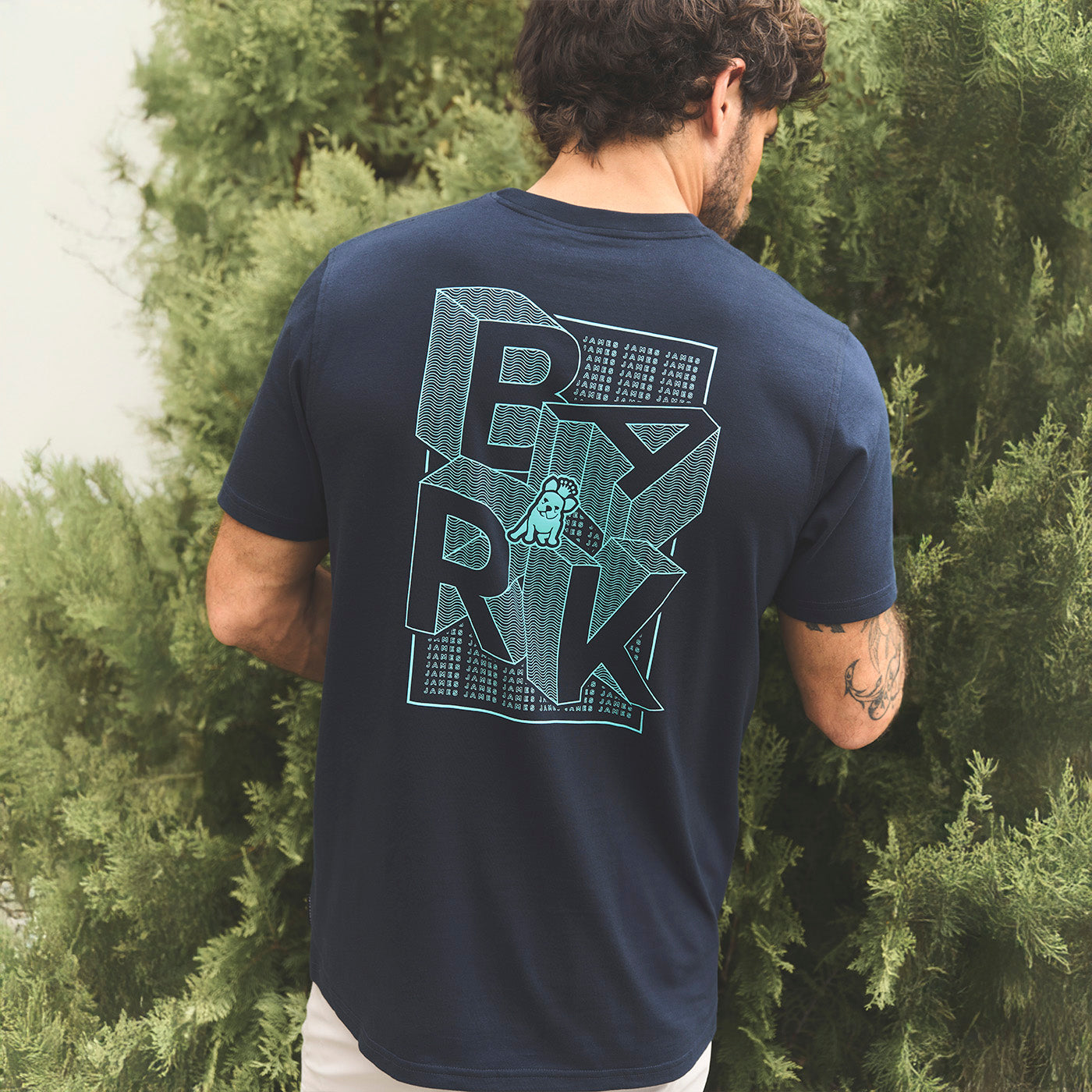 Man wearing a navy blue t-shirt with a graphic design on the back, standing in front of greenery.