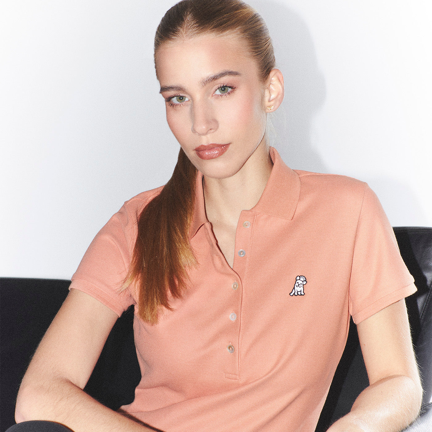 Woman wearing a pink polo shirt with a small logo, sitting on a black couch against a white background