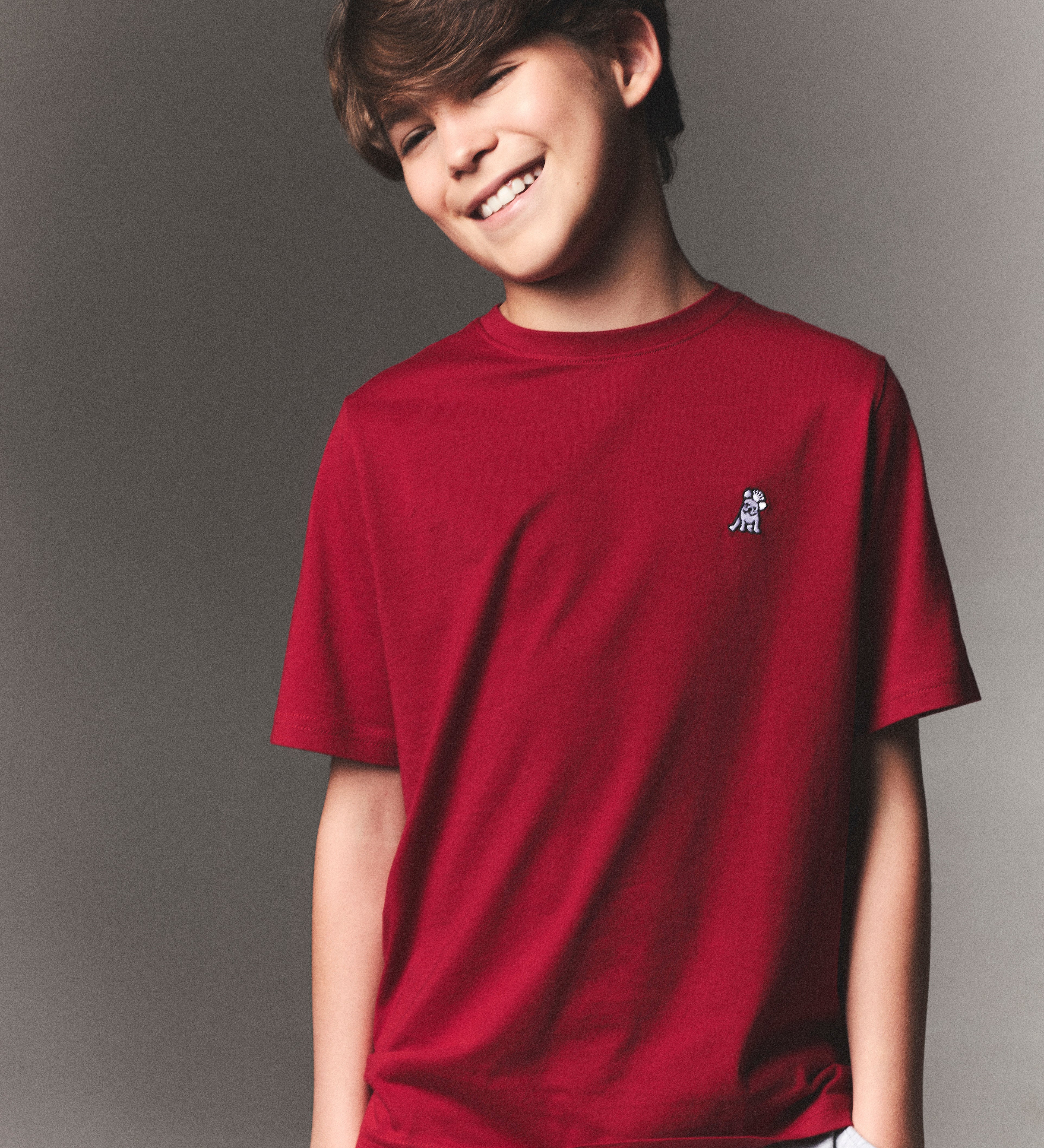 Young boy smiling in a red James Bark crew neck t-shirt with white embroidered logo, paired with light grey drawstring shorts