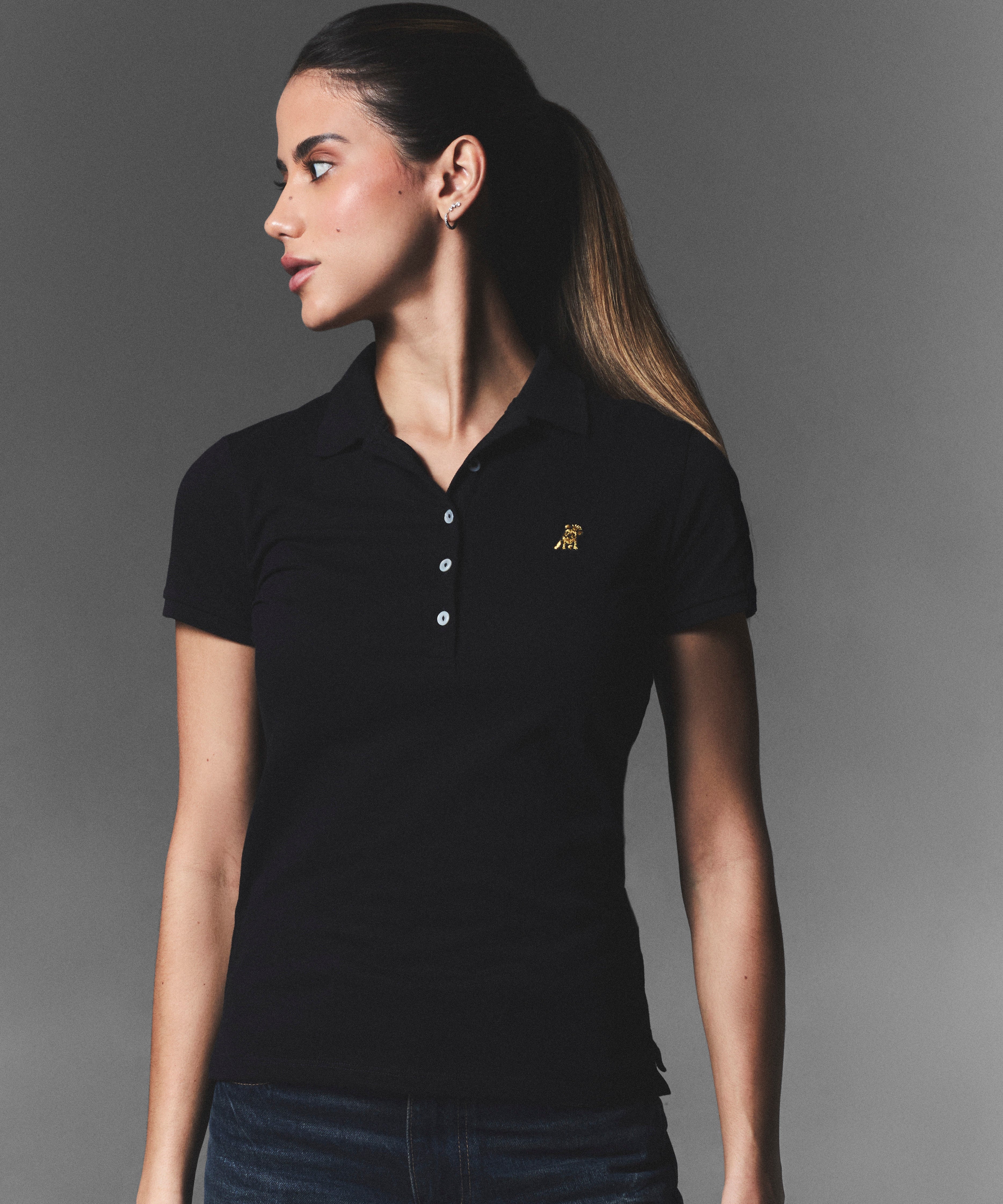 Female model wearing a fitted black James Bark polo shirt with gold embroidered logo on the chest, styled with dark denim jeans