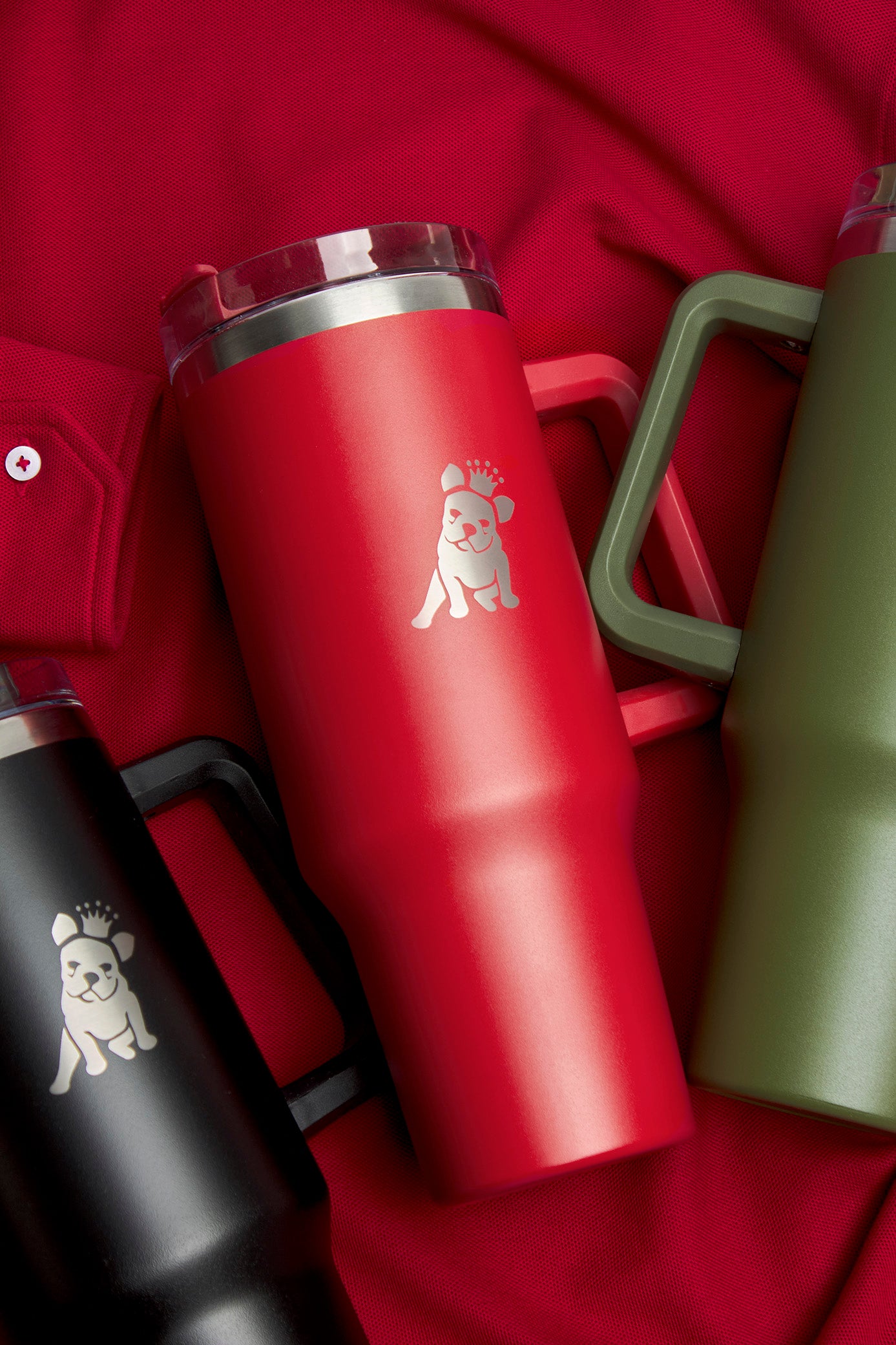 Three insulated travel mugs with handles in black, red, and green colors are arranged on a red fabric background. Each mug features a silver embossed dog logo wearing a crown, with the red mug positioned prominently in the center.