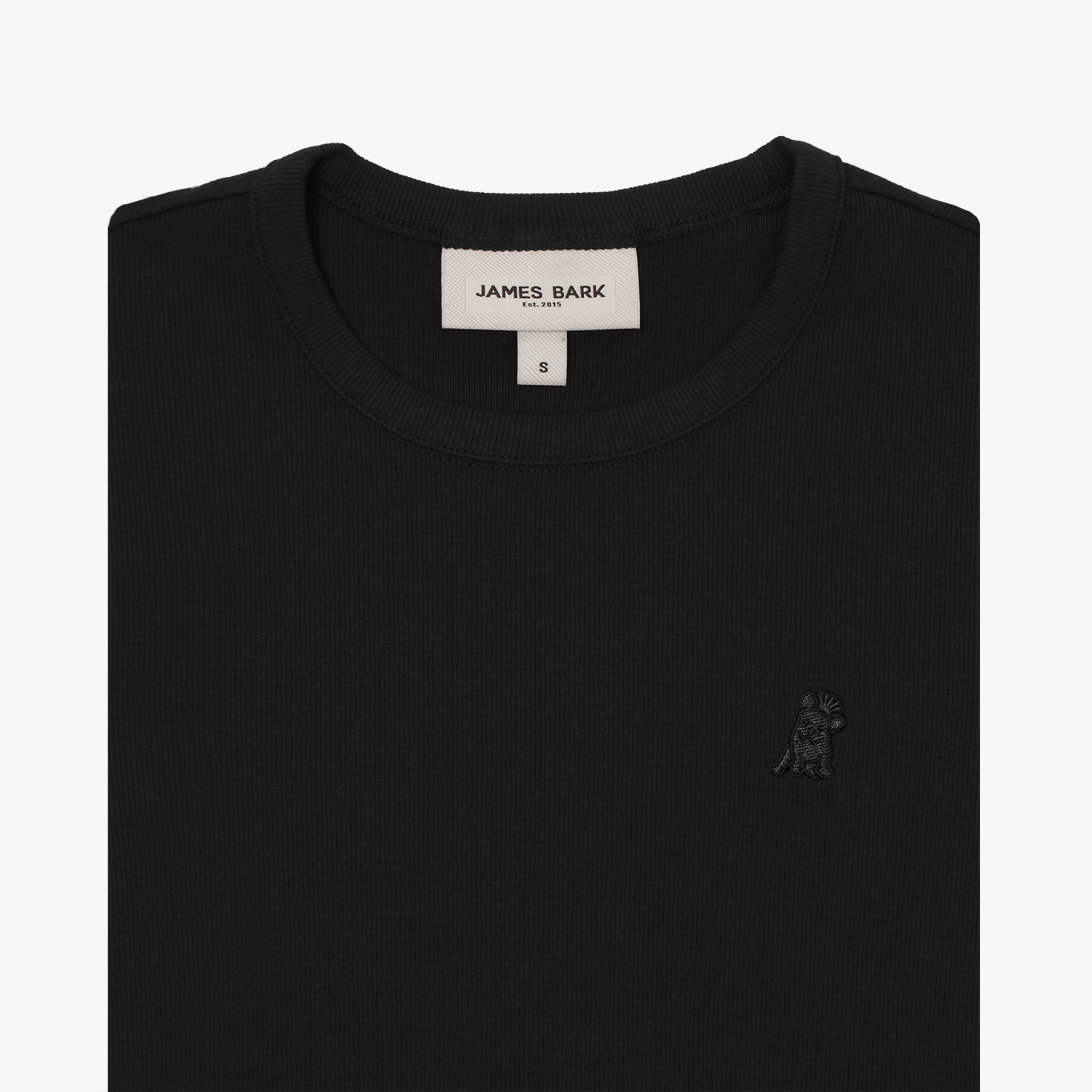 A close-up of a black short-sleeve T-shirt, neatly folded to showcase the ribbed crewneck collar and finely knit fabric texture. The white neck label displays the brand name "JAMES BARK" with "Est. 2015" beneath it, along with the size "S." A subtly embroidered logo of a dog, matching the black fabric, is positioned on the left chest area. The material appears soft and high-quality.