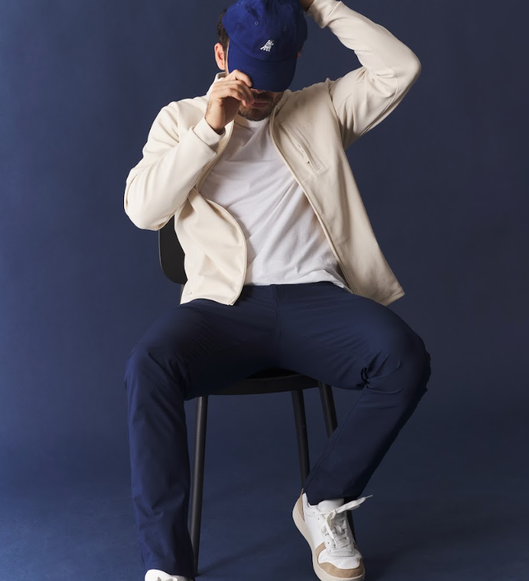 Person sitting on a chair wearing a white jacket, white shirt, navy pants, and white sneakers, adjusting a blue cap against a dark background.