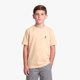 A young boy wearing a light peach T-shirt with a small logo on the left chest, paired with gray pants, standing against a white background.