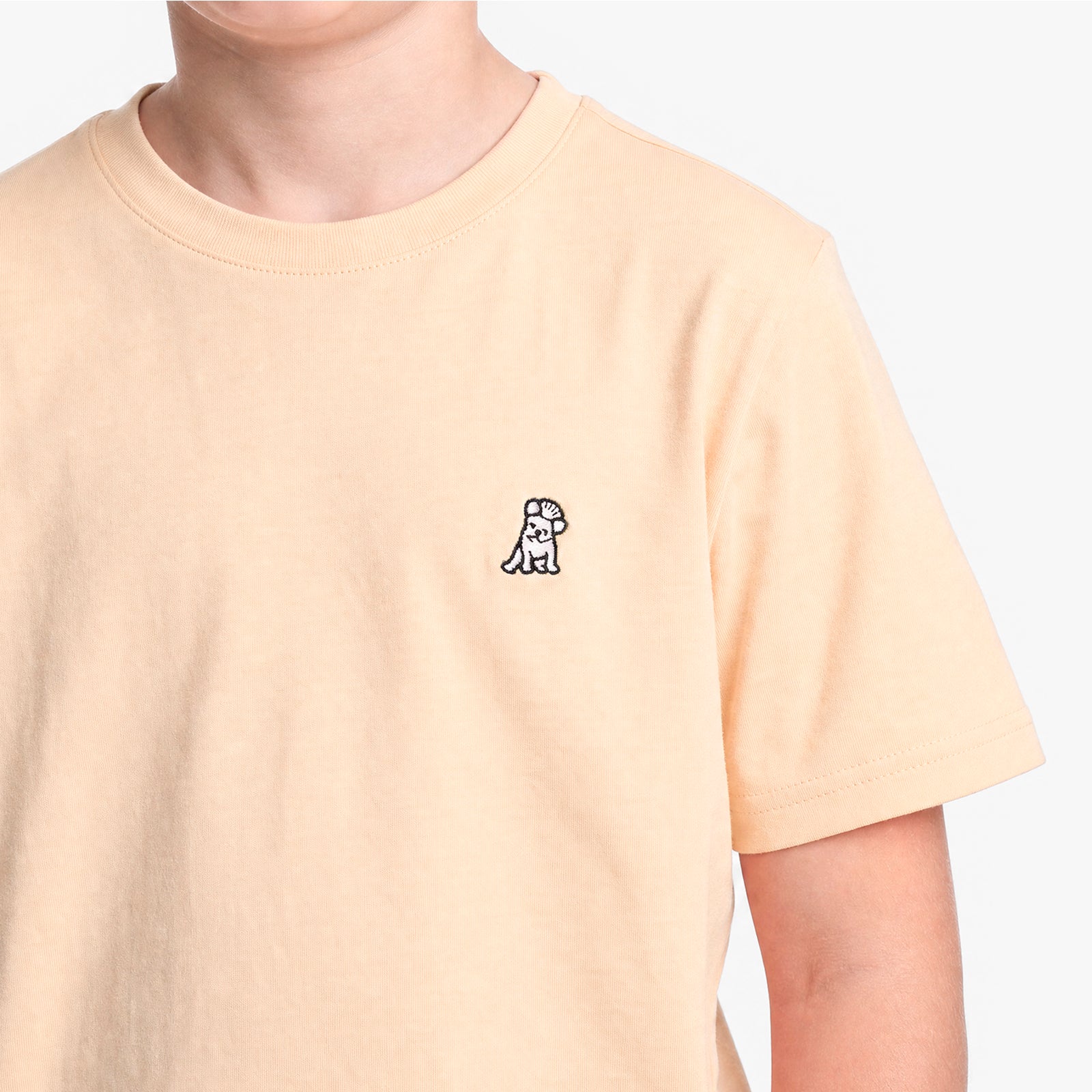 A close-up of a person wearing a light peach T-shirt with a small black and white dog on the left chest, set against a white background.