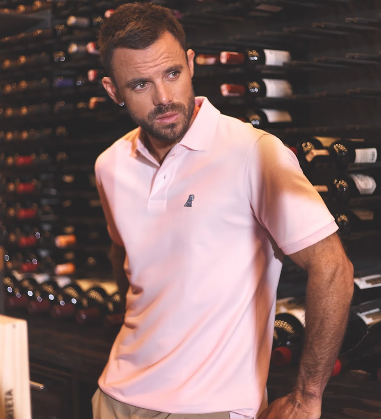 Man standing in a wine cellar wearing a light pink polo shirt with a logo.