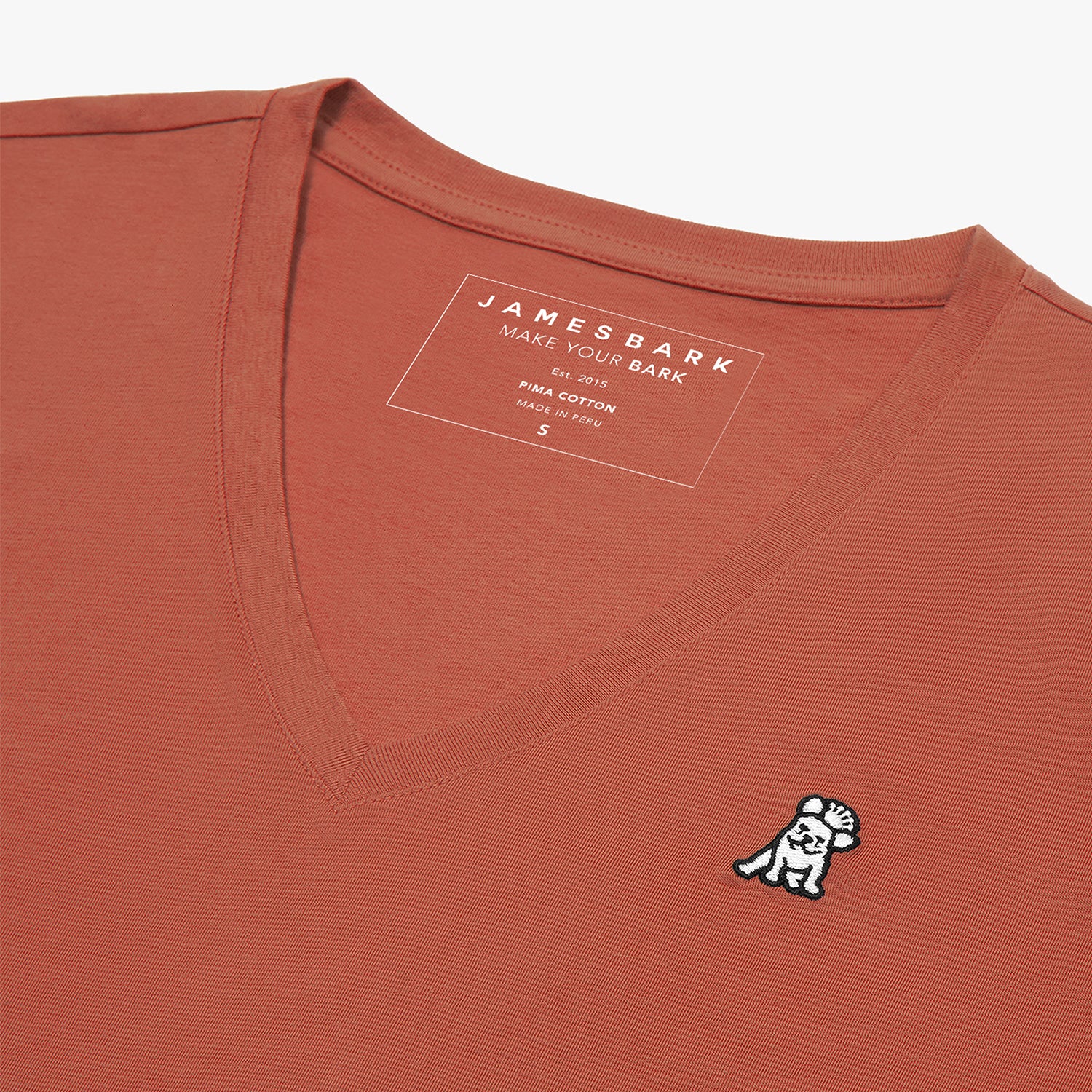 Close-up angled view of the inside collar area of the terracotta V-neck t-shirt laid flat, showing the V-neckline, the printed "JAMES BARK" brand tag, and the black and white embroidered dog logo on the chest
