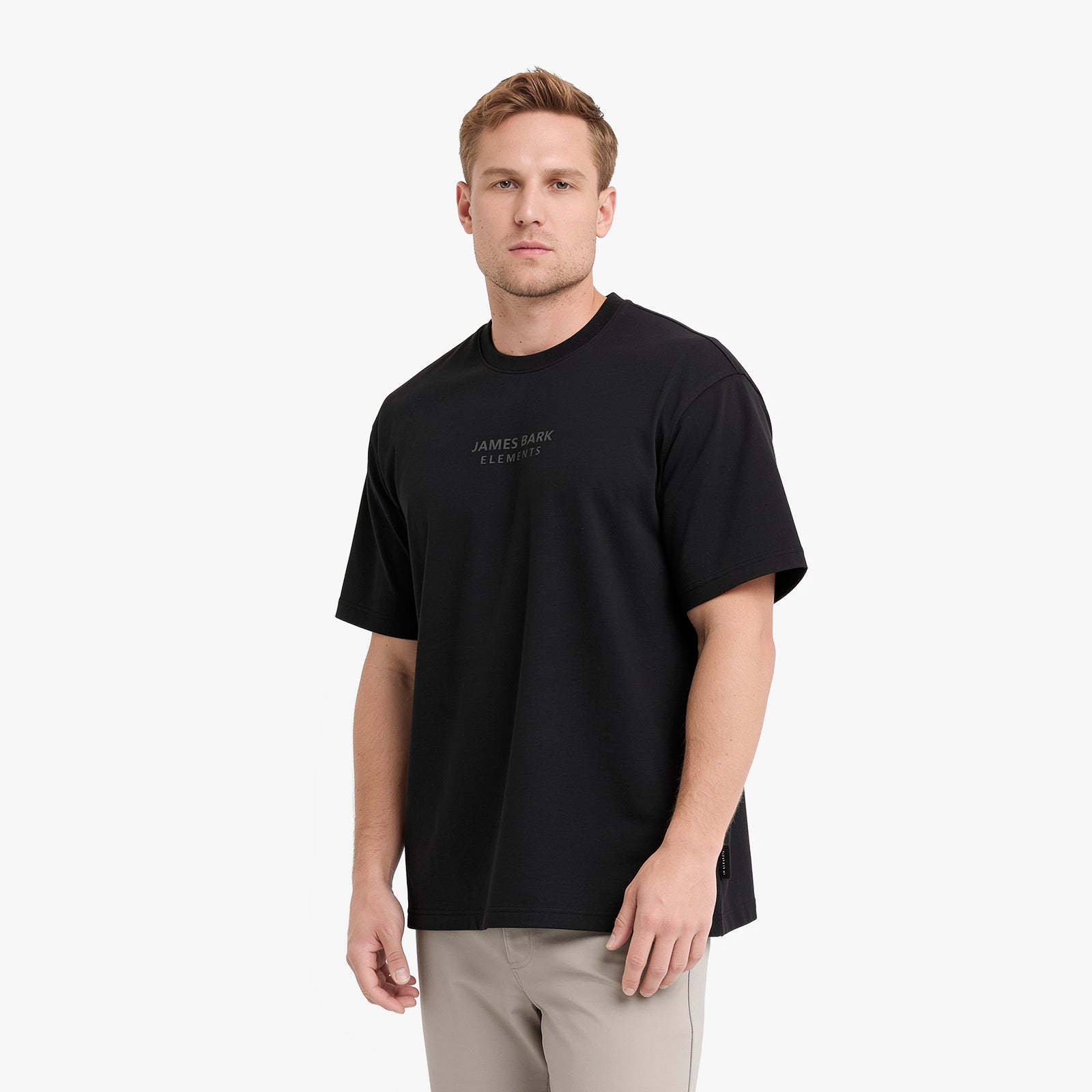 A man wearing a black crewneck t-shirt with 'ELEMENTS' printed in small gray letters on the left chest, paired with light beige trousers
