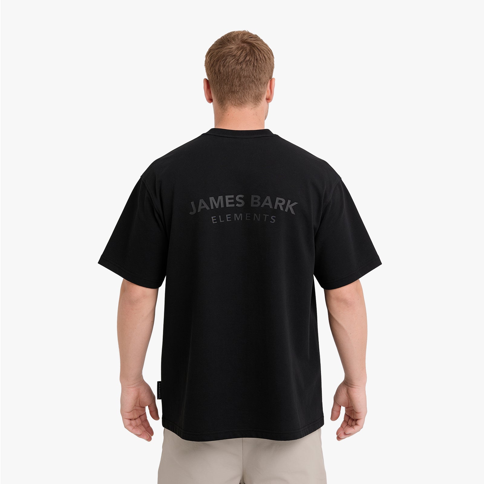 A man viewed from behind wearing a black t-shirt with 'JAMES BARK ELEMENTS' printed in a dark gray tone on the back, paired with light beige pants