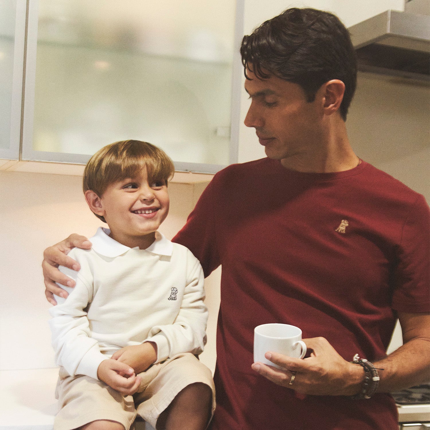 Father and son sharing a warm moment in the kitchen. The father, wearing a burgundy James Bark t-shirt, lovingly holds a coffee mug while looking at his smiling son, who is dressed in a cream sweatshirt and beige shorts with socks. A cozy and stylish family scene highlighting matching casual elegance