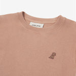 A close-up of a dusty rose-colored crewneck sweatshirt, showing the ribbed collar and finely knit fabric texture. The white neck label displays the brand name "JAMES BARK" with "Est. 2015" beneath it. A small embroidered logo of a dog in a darker brown thread is positioned on the left chest area, subtly contrasting with the fabric. The material appears soft and high-quality.