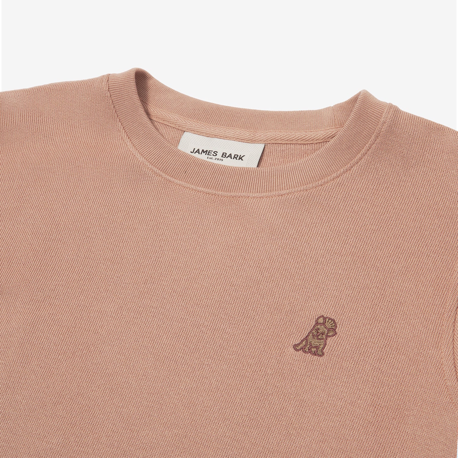 A close-up of a dusty rose-colored crewneck sweatshirt, showing the ribbed collar and finely knit fabric texture. The white neck label displays the brand name "JAMES BARK" with "Est. 2015" beneath it. A small embroidered logo of a dog in a darker brown thread is positioned on the left chest area, subtly contrasting with the fabric. The material appears soft and high-quality.