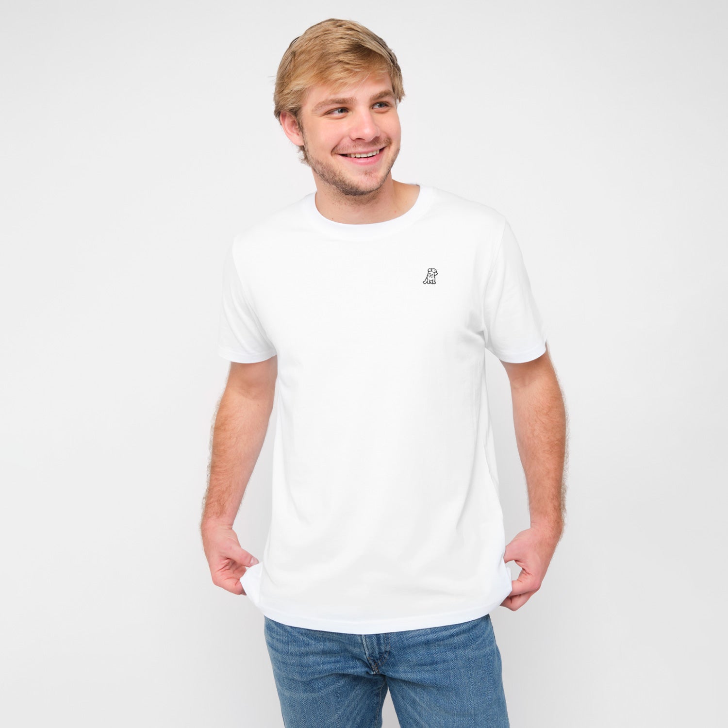 A man smiling and standing, wearing the white T-shirt with a small embroidered logo on the chest, paired with jeans.